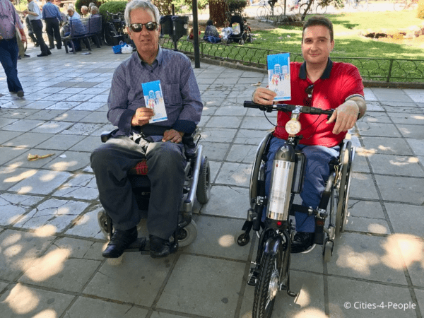Two people using Trikala's electric wheelchair scooters, holding Cities-4-People brochures