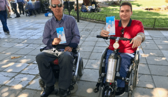 Two people using Trikala's electric wheelchair scooters, holding Cities-4-People brochures