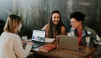 Three young people sitting in front of their laptops at a table laughing.